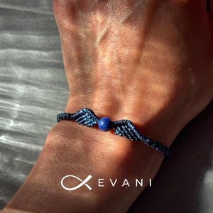 Könnte beinhalten: Ein dunkelblaues Makramee-Armband mit einer zentralen runden blauen Steinperle, die von zwei gewebten flügelförmigen Elementen flankiert wird. Das Armband ist mit einer dünnen Kordel geknüpft und zeigt den weißen Schriftzug EVANI mit einem Logo.