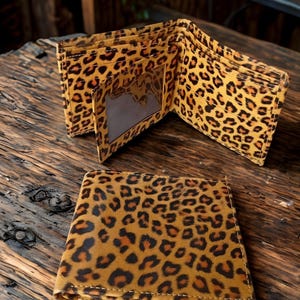 Puede incluir: Carteras con estampado de leopardo abiertas y cerradas sobre una superficie de madera. Las carteras son de color beige con manchas negras y marrones. Una cartera está abierta, revelando ranuras para tarjetas y una ventana de identificación transparente.