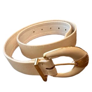 Pode incluir: Um cinto de cor creme com uma tira lisa e sólida que apresenta uma série de orifícios de ajuste. O cinto é fixado por uma fivela grande e curva de cor creme, acentuada com detalhes em metal dourado no pino e no passador.