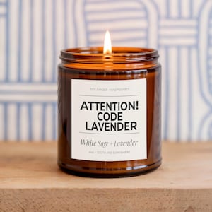 May include: A lit soy candle in a brown glass jar. The label reads "ATTENTION! CODE LAVENDER" with "White Sage + Lavender" below. The candle is 4 oz. and hand-poured. The background is a blue and white patterned wall.