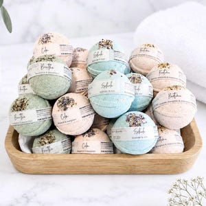 May include: A collection of spherical bath bombs in a wooden tray, featuring various pastel colours including light green, blue, and peach. Each bath bomb is wrapped with a paper label displaying text such as Breathe, Calm, and Exhale, with dried botanicals on top.