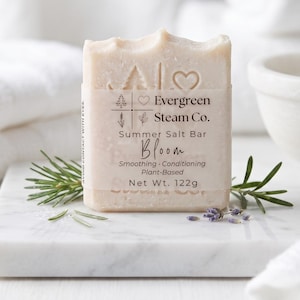 Könnte beinhalten: Ein hellfarbiger, rechteckiger Seifenblock mit einem Papieretikett mit der Aufschrift "Evergreen Steam Co. Summer Salt Bar Bloom". Das Etikett enthält auch eine Baum-, Herz- und Blattgrafik. Die Seife ist pflanzlich und wiegt 122g. In der Nähe befinden sich Zweige von Rosmarin und Lavendel.