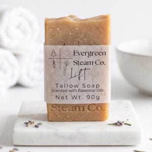 May include: A light brown tallow soap bar with a paper label that reads "Evergreen Steam Co. Lift Tallow Soap Scented with Essential Oils Net Wt. 90g." The soap is displayed on a white marble square, with lavender sprigs.