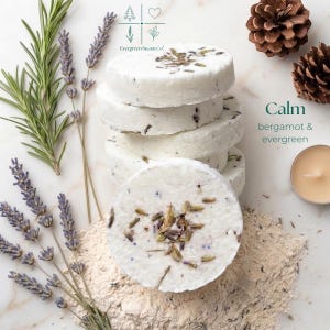 Peut inclure: Une pile de galets de douche ronds blancs parsemés de boutons de lavande séchés, avec l'inscription Evergreen Steam Co. Calm bergamot & evergreen. Ils sont entourés de brins de romarin frais, de tiges de lavande, de deux pommes de pin et d'une bougie.