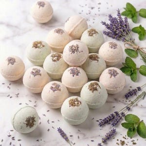 May include: A collection of round, pale-colored bath bombs arranged on a white marble surface. Each spherical bath bomb is topped with a small cluster of dried lavender buds. Sprigs of fresh lavender and green mint leaves are scattered nearby.