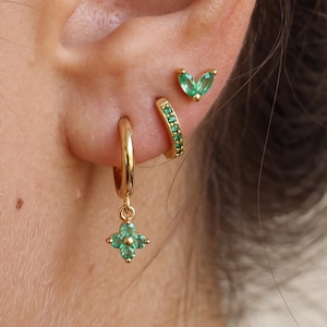 May include: A set of three gold earrings featuring green stones. The bottom earring is a small hoop with a dangling four-petal flower charm. The middle earring is a small hoop with embedded green stones. The top earring is a two-stone leaf stud.