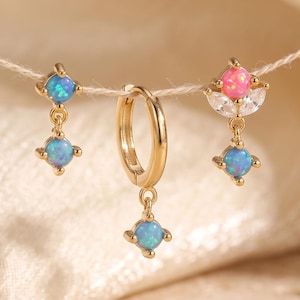 May include: Three gold-toned earrings suspended on a string. The left earring features two round blue opalescent stones. The centre earring is a small hoop with a single blue stone drop. The right earring has a pink stone and clear crystals.
