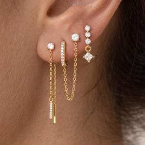 May include: A set of four gold-toned earrings featuring clear stones. One earring has a dangling chain with a bar, one is a small hoop, one has a chain connecting two points, and one features a vertical row of stones with a dangling square stone.