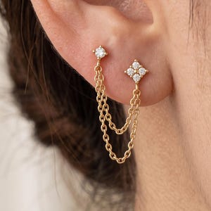 May include: A gold double chain earring featuring two studs connected by two delicate gold chains. One stud is a single round clear stone, while the other is a four-stone cluster forming a diamond shape. The earring is worn in two piercings.