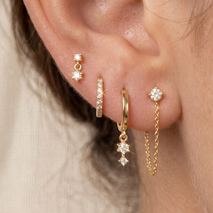 May include: Close-up of a person's ear adorned with multiple gold earrings. The earrings include a small hoop, a stud with a dangling charm, a stud with a chain, and a stud with a small dangling earring. All earrings are embellished with small, clear gemstones.