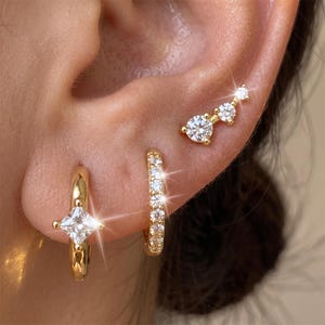 May include: Three gold-tone earrings featuring clear, sparkling stones. One hoop earring has a single square-cut stone, another is a thin hoop lined with small round stones, and the third is a curved stud with three graduated round stones.