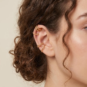May include: A close-up of a person's ear adorned with multiple gold ear cuffs and a hoop earring. The ear cuffs feature various designs, including a triple band, a twisted rope, and a chain link. The jewelry is shiny and reflective.