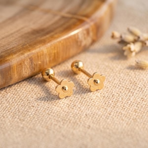 Op de afbeelding: Een paar goudkleurige bloemvormige oorstekers. Elke oorbel heeft een klein, vijfbladig bloemontwerp met een afgeronde pin en een bolvormige achterkant. De oorbellen worden gepresenteerd op een getextureerd, beige oppervlak.