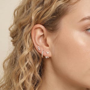 May include: An ear featuring several gold-toned earrings. There are small stud earrings with clear gemstones, a small hoop earring also with gemstones, and leaf-shaped earrings with clear gemstones. The earrings are delicate and elegant.