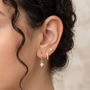 May include: A set of gold earrings with a diamond-shaped charm, a small diamond stud, and a hoop earring with small, clear stones. The earrings are displayed on a person's ear, showcasing their design and sparkle.