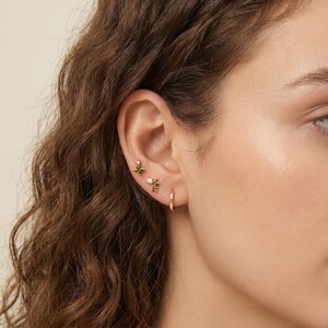 May include: A gold ear stack featuring three distinct pieces: a small stud with a green gemstone, a second stud with a small clear stone and green accents, and a thin gold hoop earring. The jewelry is worn in the earlobe and cartilage area.