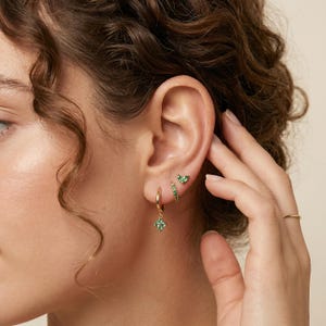 Puede incluir: Pendientes dorados con piedras preciosas verdes. Los pendientes incluyen un aro pequeño con un colgante cuadrado, un aro pequeño con piedras verdes y un pendiente de botón con un diseño de hoja. También se ve un anillo de oro.