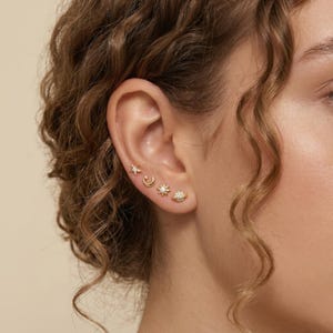 May include: A person's ear is shown with four gold stud earrings. The earrings are shaped like a star, a crescent moon, a sun, and a planet. Each earring has small, sparkling details, giving a delicate and elegant appearance. The earrings are a celestial theme.