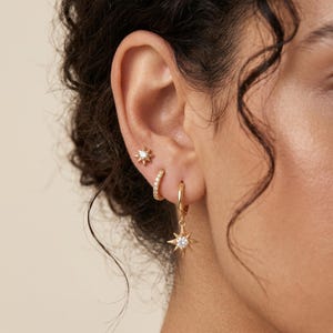 May include: Gold-toned earrings displayed on an ear. The set includes a starburst stud, a small hoop with clear stones, and a larger hoop with a dangling starburst pendant with a clear stone center. The earrings are set against a neutral background.