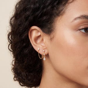 May include: Gold-toned double earrings with a delicate chain. The earrings feature small, round, and square-cut clear gemstones. The design includes a stud earring and a dangling earring connected by a chain.
