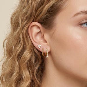 May include: A close-up of a person's ear adorned with three gold earrings. The top earring is a small triangle shape with three clear stones. The middle earring is a small gold hoop. The bottom earring is a gold hoop with clear stones.