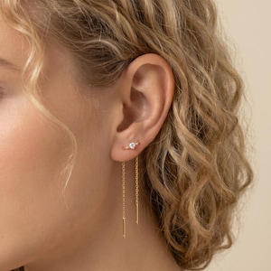 Könnte beinhalten: Goldene Ohrringe mit einem kleinen, klaren Edelstein-Stecker und einer zarten Kette. Die Kette hängt nach unten und endet in einem kleinen Goldstab. Die Ohrringe werden an einem Ohr gezeigt.