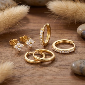 May include: A selection of gold earrings, including stud earrings with a floral design, small hoops, and larger hoop earrings. The earrings are embellished with small, clear gemstones. The jewellery is displayed on a wooden surface.