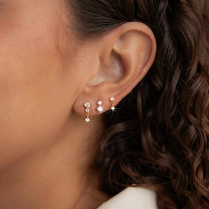May include: Close-up of three sets of gold earrings with clear, sparkling gemstones. The earrings are in different styles, including studs and dangling designs. The earrings are worn on an ear, showcasing the jewelry's delicate details and shine.