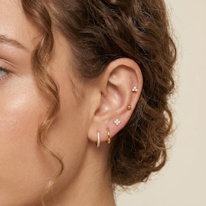 May include: Close-up of an ear adorned with multiple gold earrings. The ear features a small hoop earring with embedded crystals, a plain gold hoop, and several stud earrings, including a ball, a cluster of three small stones, and a four-stone flower design.