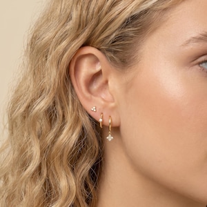 May include: Close-up of a person's ear adorned with three different gold earrings. The ear has a small stud earring with three small white gems, a small hoop earring with a textured design, and a larger hoop earring with a dangling four-petal flower design.