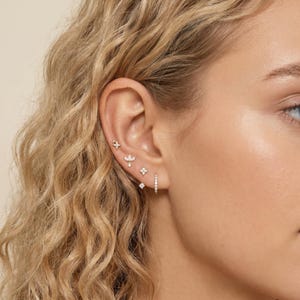 May include: A collection of gold earrings, including a star, floral, and square design, and a small hoop earring. The earrings are adorned with small, sparkling gemstones. The earrings are displayed on an ear.