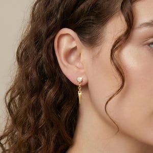 May include: A gold-colored earring with a small, round, clear gemstone at the top. The earring has a dangling design with a small, pointed, gold-colored spike. The earring is worn on a person's ear.