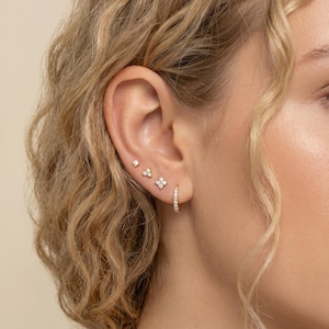 May include: An ear featuring a collection of gold earrings. The earrings include a small square stud, a cluster of three small stones, a four-petal flower design, and a small hoop earring with multiple small stones.