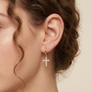 May include: A gold-colored hoop earring with a dangling cross pendant. The cross is encrusted with small, clear gemstones. The earring is shown on a person's ear, with their hair and shoulder visible in the background.