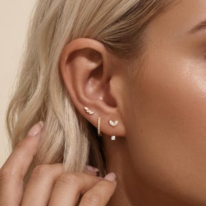 May include: A set of three gold earrings worn in one ear. The top earring is a small leaf-shaped stud with three clear stones. The middle earring is a thin gold hoop with pavé stones. The bottom earring is a V-shaped stud with two clear stones.