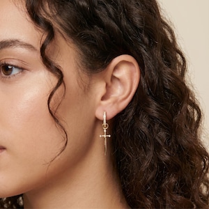 May include: A gold hoop earring featuring a small, dangling sword-shaped charm. The hoop is encrusted with tiny clear stones, and the sword charm includes a cross-guard detail with a small embedded stone at its center, creating a delicate look.