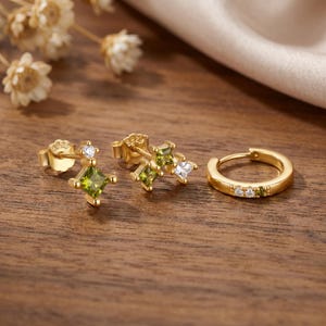 May include: A set of three gold-tone earrings including two stud earrings with square-cut olive green and clear stones, and one small hoop earring featuring a row of alternating clear and olive green stones, all marked with S925 stamp.