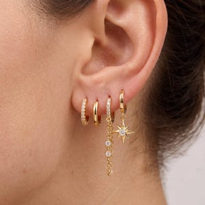 May include: A set of four gold-tone hoop earrings worn in a single ear. The first is a small pavé hoop, the second a plain polished hoop, the third a hoop with a dangling chain featuring three round crystals, and the fourth a star-shaped charm.
