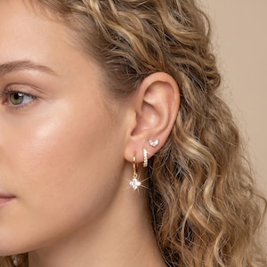 May include: A close-up of an ear adorned with multiple gold earrings. The ear features a small, curved stud, a small hoop with embedded crystals, and a larger hoop with a dangling, four-petal flower design, all in gold with clear crystals.