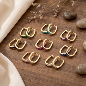 May include: A selection of gold-plated hoop earrings, each set with different coloured gemstones. The earrings feature green, turquoise, purple, black, pink, blue, and clear stones. They are arranged on a wooden surface.