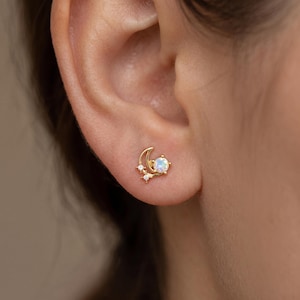 May include: A gold stud earring featuring a crescent moon shape, a round iridescent opal stone, and two small clear accent stones. The earring is positioned on the earlobe, showcasing a celestial design with a polished gold metal finish.