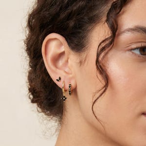 May include: A close-up of a person's ear adorned with multiple earrings. The earrings include small black gemstone studs, gold hoop earrings with black stones, and gold hoop earrings with a black flower pendant. The earrings are set against the backdrop of the person's skin and hair.