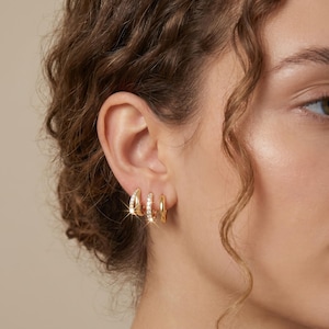 May include: Three pairs of gold hoop earrings are displayed on an ear. The first pair is a small, thick hoop. The second pair is a small hoop with clear, sparkling stones. The third pair is a small, thick hoop.