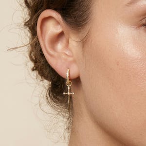 May include: A gold-colored hoop earring with small, clear stones encircling the hoop. A gold sword pendant with a cross-shaped hilt and a pointed blade hangs from the hoop. The earring is worn on a person's ear.