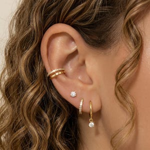 May include: A collection of gold earrings and ear cuff. The ear cuff features two rows of small, sparkling stones. There is a small stud earring, a hoop earring with a dangling stone, and a small hoop earring with stones.