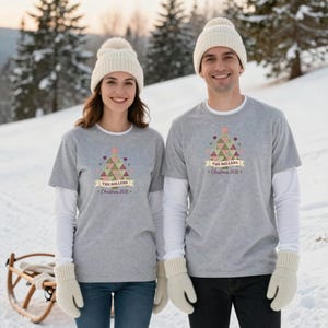 May include: Two grey short-sleeve t-shirts worn over white long-sleeve shirts, paired with cream-colored knit beanies and mittens. Each shirt features a graphic of a patterned Christmas tree with the text THE MILLERS Christmas 2026.