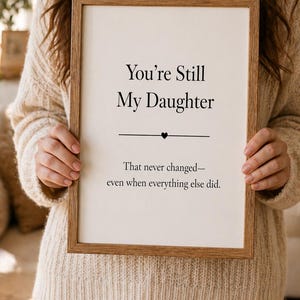 May include: A framed print with the text "You're Still My Daughter" in a black font. Below the text is a horizontal line with a heart symbol. The bottom text reads "That never changed—even when everything else did."
