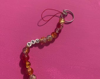 May include: A beaded keychain with the letters "ASU" on white letter beads. The keychain features a variety of colorful beads in shades of red, orange, and clear. A silver-colored ring and red string complete the design.