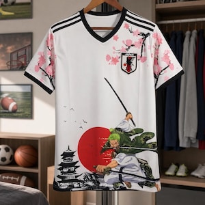 May include: A white V-neck t-shirt featuring black shoulder stripes, pink cherry blossom branches, and a Japan crest. The front displays a graphic of a green-haired swordsman with three swords, a red sun, and a black pagoda silhouette.