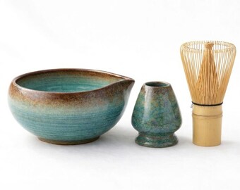 May include: A matcha tea set featuring a turquoise and brown ceramic bowl with a spout, a small ceramic tea bowl, and a bamboo whisk. The set is designed for preparing traditional matcha tea.